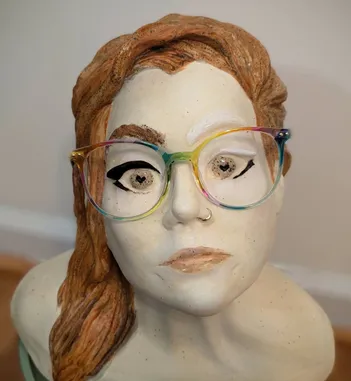 A photo of the artist - a redhead with rainbow glasses against a blue and gold background